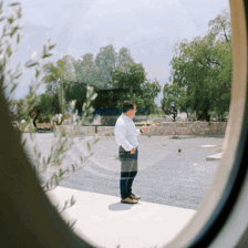 Person in white shirt seen through round window, standing outdoors near trees and stone wall.
